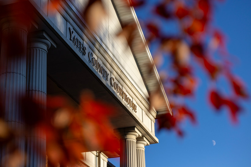 An image of the Yates County Courthouse. In the foreground you can see blurred fall leaves that frame the courthouse, creating a dramatic effect.