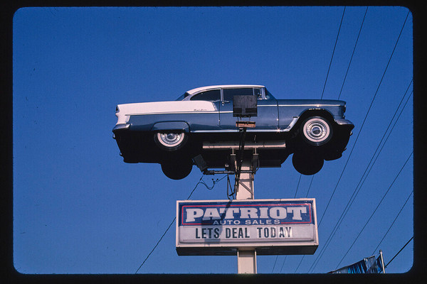 A blue car suspended on a post against a blue sky with a sign below reading "PATRIOT AUTO SALES: LETS DEAL TODAY"