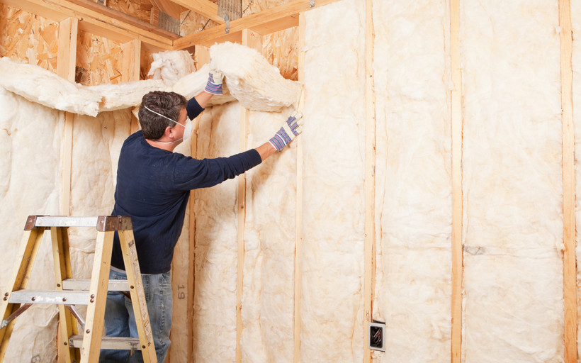 A man wearing a mask installs pink fiberglass insulation on the inside of a home.