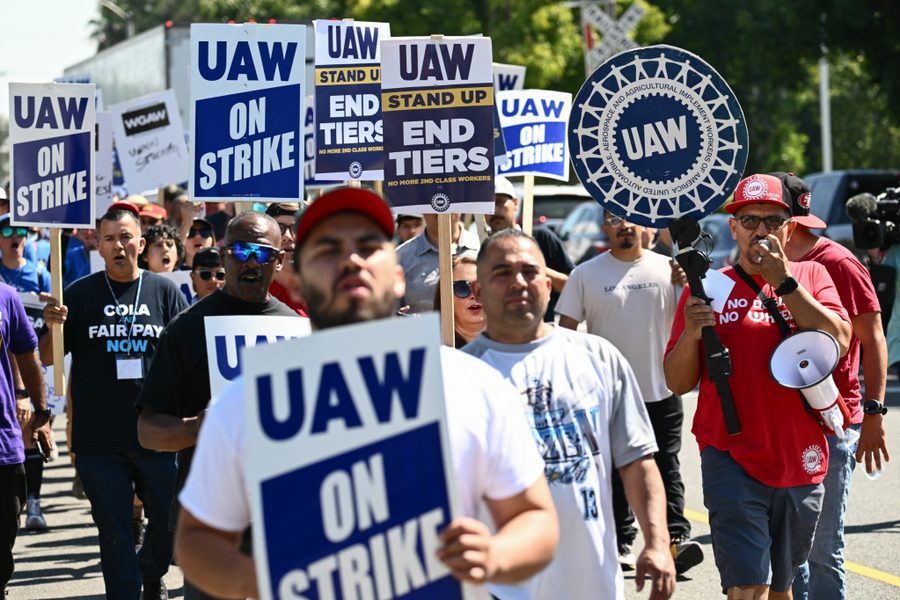 A group of people march down a street, one of them speaking into a megaphone. They hold sings that read, "UAW on strike" and "UAW: Stand up, en tiers, no more 2nd class workers."