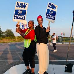 Two smiling workers stand back-to-back with signs reading, "UAW on Strike."