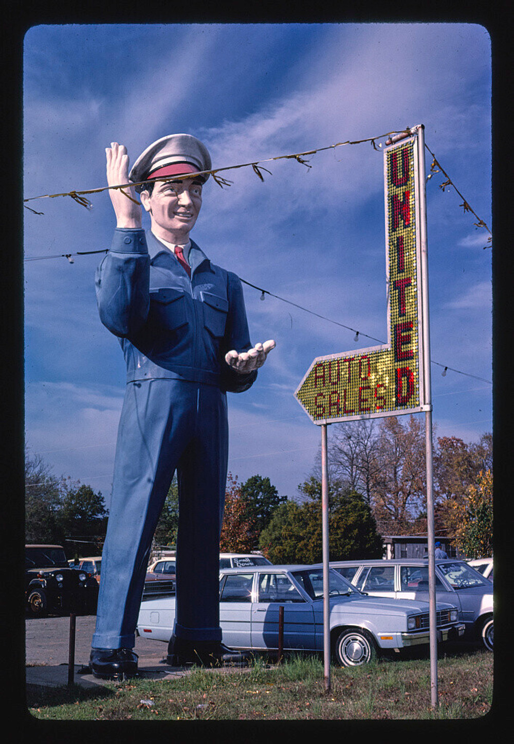 a giant statue of a mechanic in a blue suit and white hat next to an arrow-shaped sign reading UNITED AUTO SALES