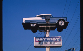 A blue car suspended on a post against a blue sky with a sign below reading "PATRIOT AUTO SALES: LETS DEAL TODAY"