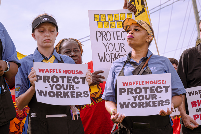 Two defiant workers stand with signs reading "Waffle House, Protect Your Workers"