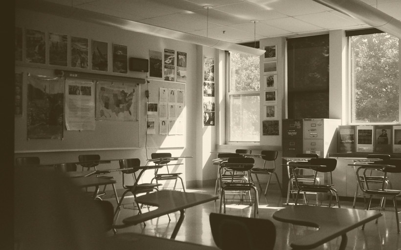 An image of a classroom with two windows and many empty desks.