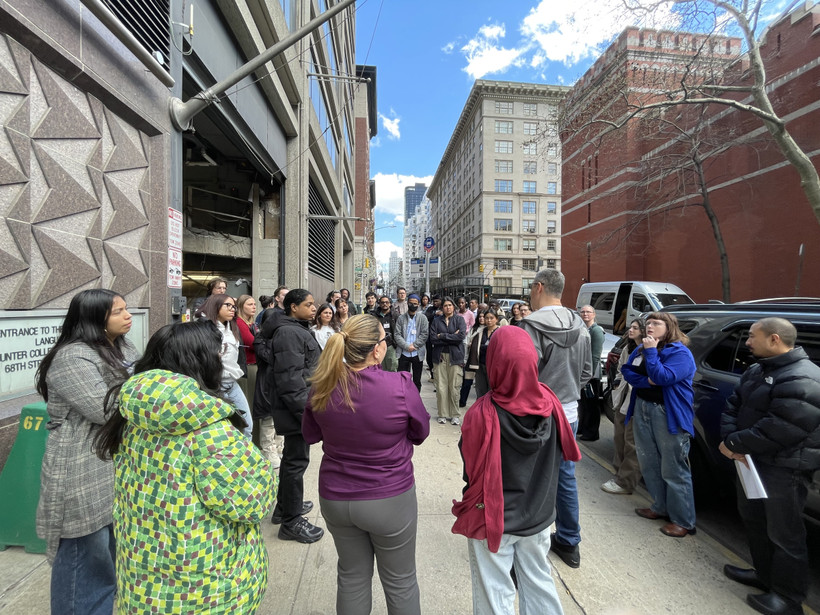 A middle-aged man speaks to an engaged group of college-age people on the sidewalk, signs showing Hunter College, 68th St, and the M66 bus are visible.