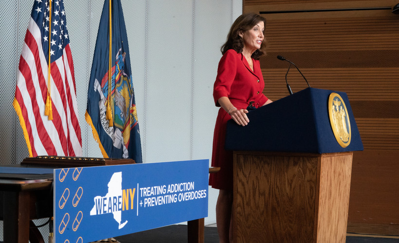 New York Governor Kathy Hochul stands at a podium next to a sign reading "treating addiction and preventing overdoses."