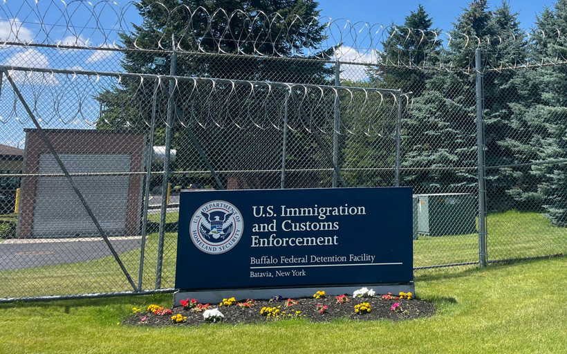 A blue sign reading "U.S. Immigration and Customs Enforcement, Buffalo Detention Center, Batavia New York" sits on the grass in front of a tall metal fence with barbed wire at the top.