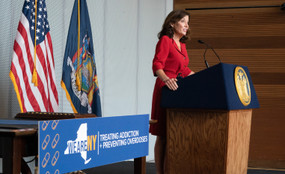 New York Governor Kathy Hochul stands at a podium next to a sign reading "treating addiction and preventing overdoses."