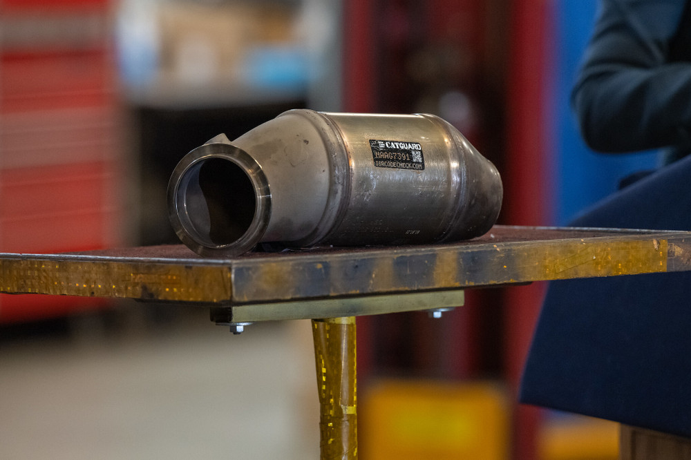 A lone catalytic converter sits displayed on a prop table. The edge of a podium and a woman's elbow are visible on the right. Elbow belongs to New York Governor Kathy Hochul.