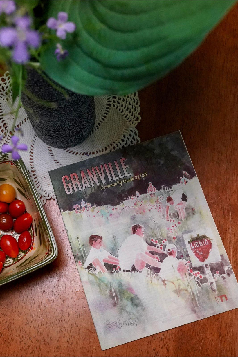 On a wooden table, a pamphlet titled "Granville Community Guide 07/08" shows a painting of people picking fruit at Liebig's Farm. Next to the pamphlet, a bowl of cherry tomatoes and a vase of flowers.
