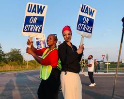Two smiling workers stand back-to-back with signs reading, "UAW on Strike."