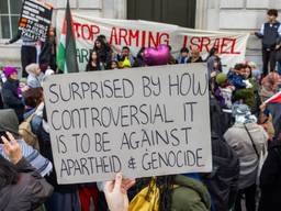 A group of people with banners and Palestinian flags stand outside a building in London. In the center of the photo, someone holds a sign that says, "Surprised by how controversial it is to be against apartheid and genocide."A banner in the background reads, in part, "Stop arming Israel."