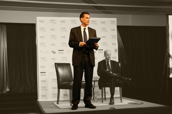 John Kaehny stands on a stage with a folder in his hands.
