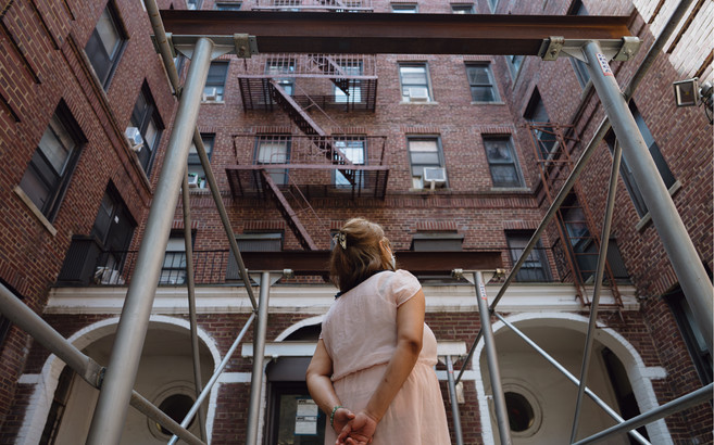 A woman stands outside a brick building, looking away from the camera, with her hands clasped behind her back.