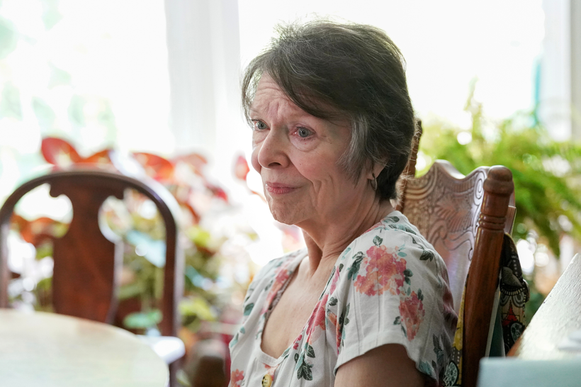 Sally Giambatista looks to the side of the camera while sitting at a dining room table. It's unclear if her expression is smiling or pained.
