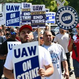 A group of people march down a street, one of them speaking into a megaphone. They hold sings that read, "UAW on strike" and "UAW: Stand up, en tiers, no more 2nd class workers."