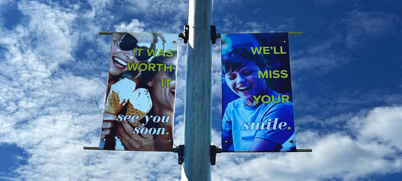Two-part banner on a light post reading "It was worth it. See you soon," and "We'll miss your smile" in front of a blue sky.