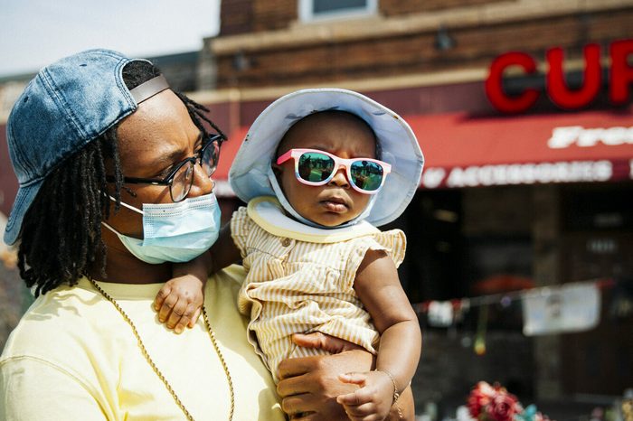 A person in a mask holds a child in sunglasses in front of the Cup Foods memorial for George Floyd. The two are wearing matching yellow clothes and blue hats.
