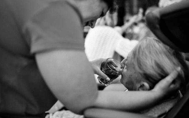 A standing woman wearing scrubs gently cradles a seated older woman's head while she helps her drink from a glass.