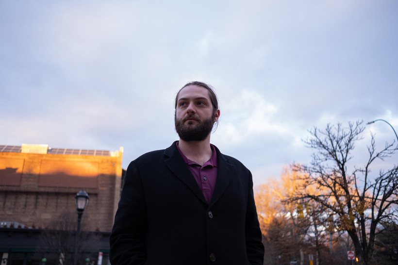 A white man with a beard stands in front of a sunlit building with a cloudy sky overhead in New York City.