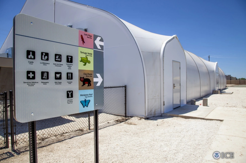 A white prefabricated structure in a desert-like area. In the foreground, a sign lists directions to a chapel, library, classrooms, grocery store, and barber; and lists the names of a few different animals, in English and Spanish.