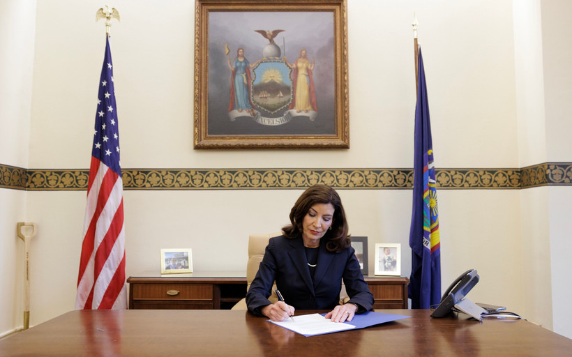 New York Governor Kathy Hochul sits at a large wooden table and signs papers in Albany, New York.