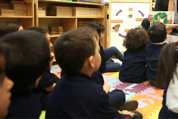 Preschool-aged children sit facing away from the camera on a colorful puzzle mat of numbers, while an adult at the front of the room shows them a page from The Very Hungry Caterpillar.