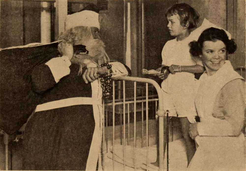 A man dressed as Santa Claus addresses a child standing on a bed in a sepia photograph of an old hospital