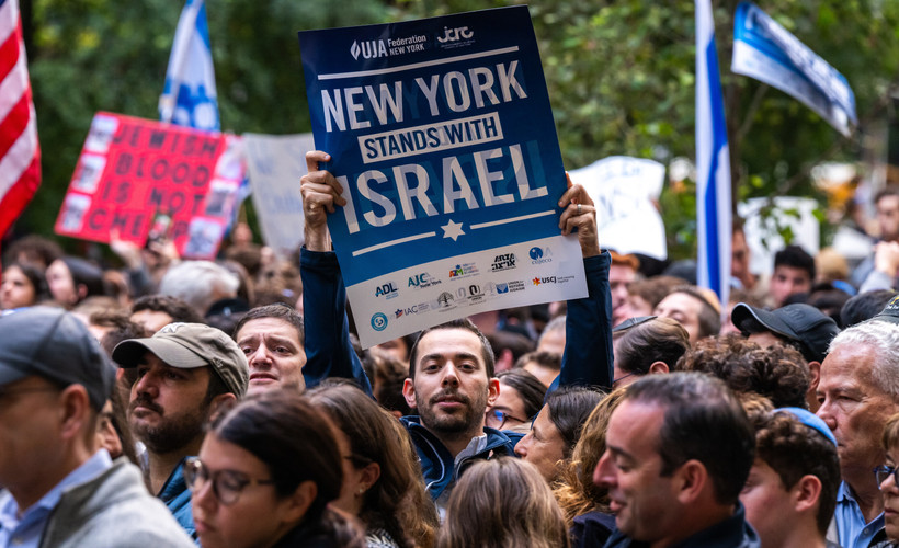 A man in a crowd holds a sign reading "New York Stands With Israel" and bearing the logos of several nonprofit groups.
