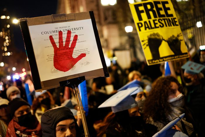 Photo of a closeup of protest signs amongst a large crowd of protestors, outside at night. The focus is on a sign that has a Columbia university graduation certificate with a red handprint on top. In the background, a yellow sign reads "FREE PALESTINE."