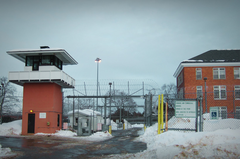 A photo of Marcy Correctional Facility shows a checkpoint for entering cars at a tall chainlink fence with barbed wire at the top. The area is snowy.