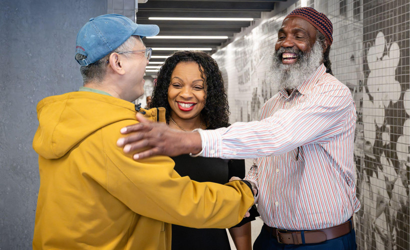 Three smiling people embrace collegially in an office hallway.