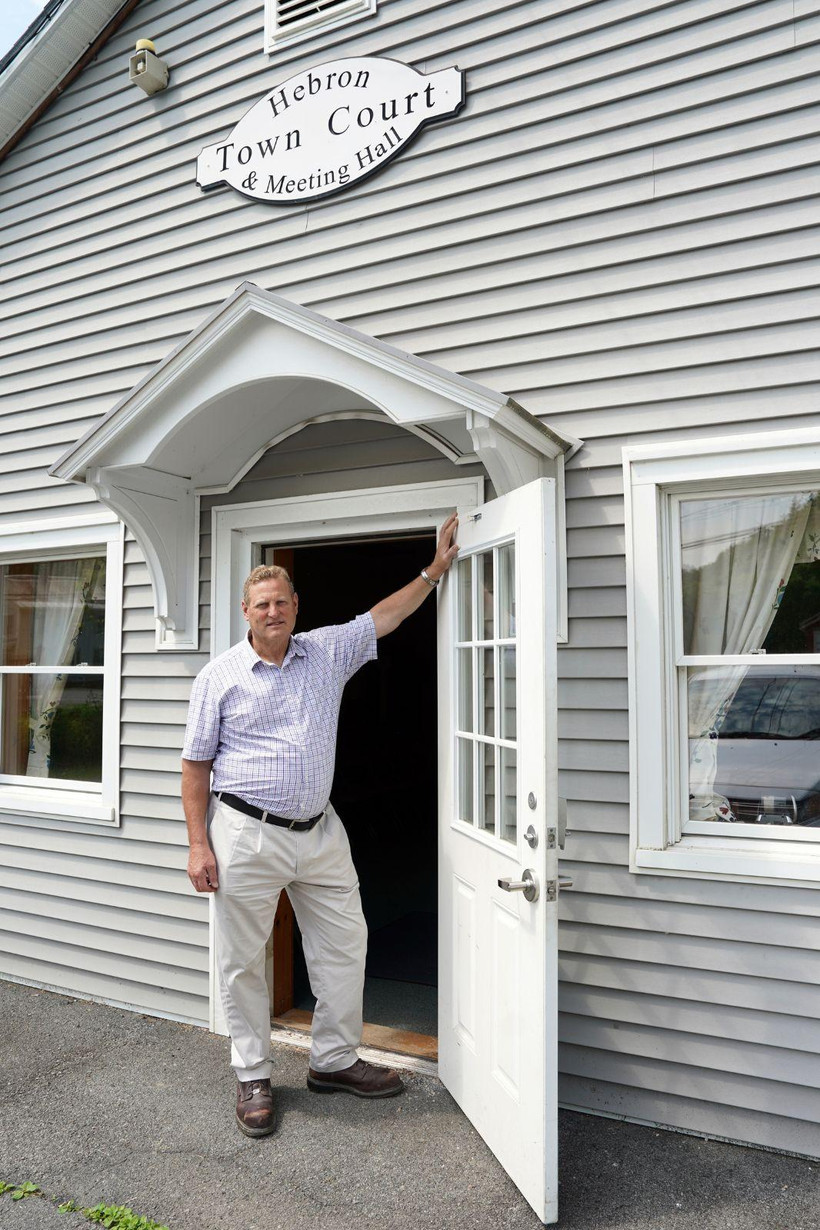 A middle-aged man wearing khakis and a short-sleeve button up shirt stands in the doorway of the Hebron Town Court and Meeting Hall, one hand up on the doorframe.
