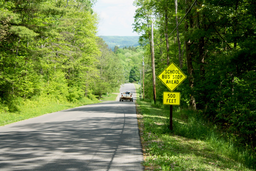A truck driving down a rural road through the woods. In the foreground, a yellow sign reads "SCHOOL BUS STOP AHEAD 500 FEET"