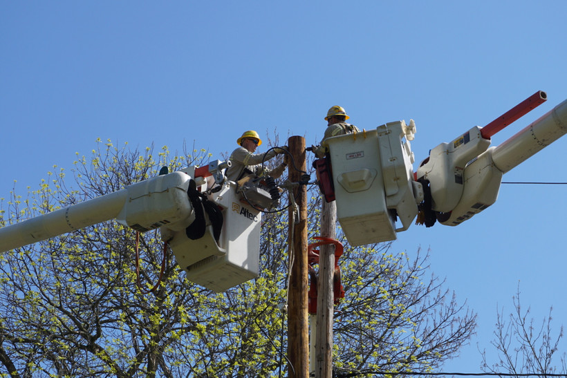 Two men in hard hats work on a utility pole from cherry picker buckets in Massena, New York.
