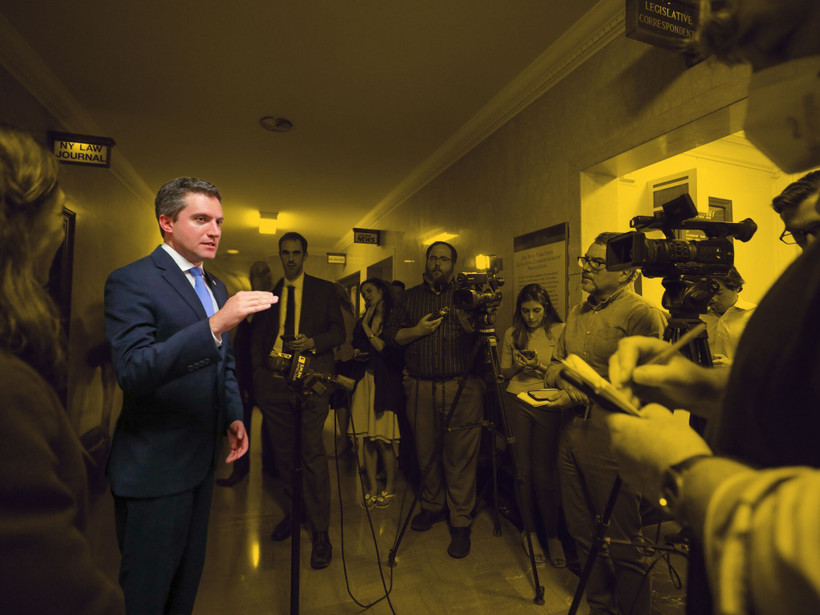 Senator James Skoufis is shown speaking to a group of reporters in a hallway.