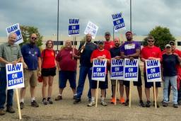 A dozen workers stand in a line with red shirts and UAW strike signs.