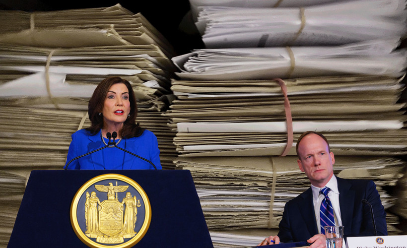 New York Governor Kathy Hochul at a podium next to her budget director, Blake Washington, with a binder, both superimposed over a photo of two stacks of paper files.