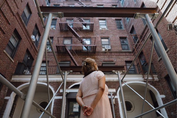 A woman stands outside a brick building, looking away from the camera, with her hands clasped behind her back.