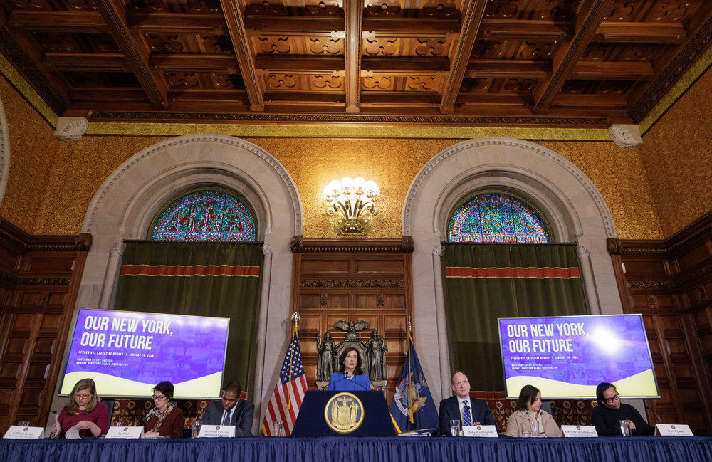 Governor Kathy Hochul stands at a podium, flanked by staff, in a room with two high, arched windows and two screens that read "Our New York, Our Future."