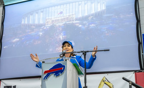 Governor Kathy Hochul of New York stands behind a podium with a Buffalo Bills logo and her arms spread wide.
