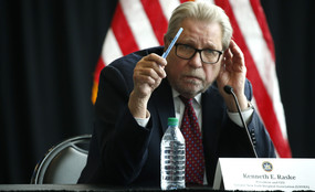 Greater New York Hospital Association President Kenneth Raske sits at a table and gestures with a pen in front of an American flag.