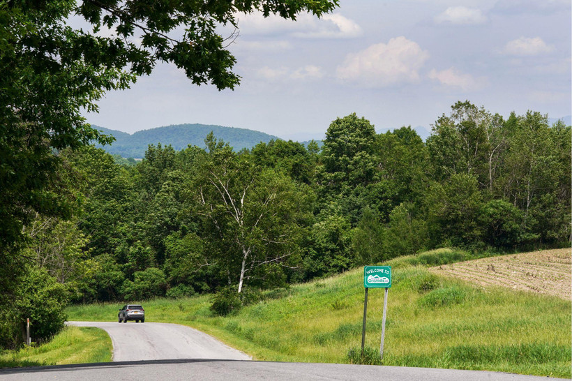 A road with a single car cuts through a grassy hillside surrounded by tres, mountains are visible in the distance. A sign reads, "Welcome to Town of Granville, NY."