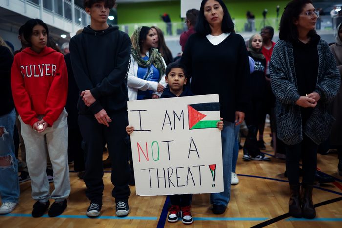 A group of people stand on a wooden floor. In the middle, a small boy is holding a banner with the Palestinian flag that reads, "I AM NOT A THREAT!"