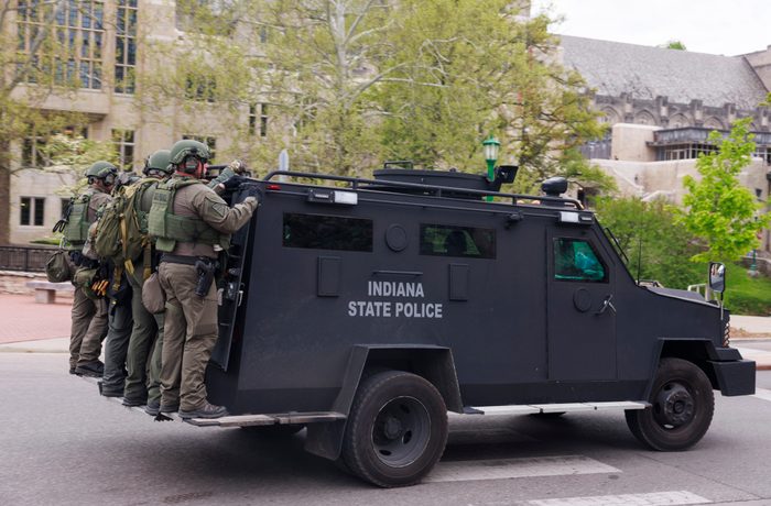 Four police officers dressed in riot gear hang onto the back of a black armored personnel carrier as it drives past university buildings.