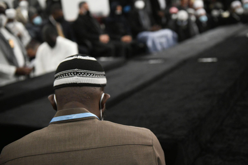 Close-up photo of a man from behind wearing a suit, kufi, and mask. In the background, many other people attending a funeral.