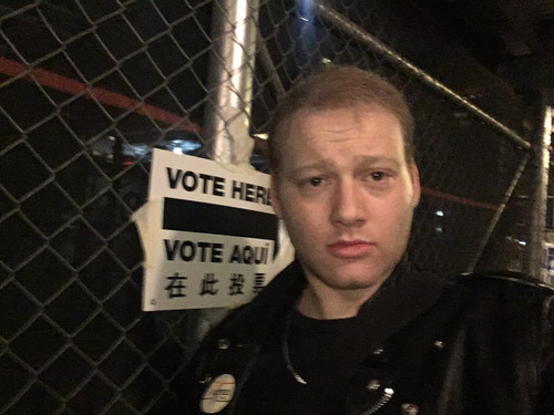 Will Bredderman stands in front of sign reading Vote Here / Vote Aquí and a chainlink fence