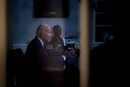 President Biden sits at a desk in profile behind a translucent glass door.