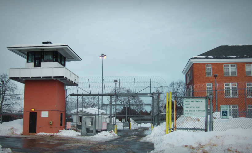 A photo of Marcy Correctional Facility shows a checkpoint for entering cars at a tall chainlink fence with barbed wire at the top. The area is snowy.